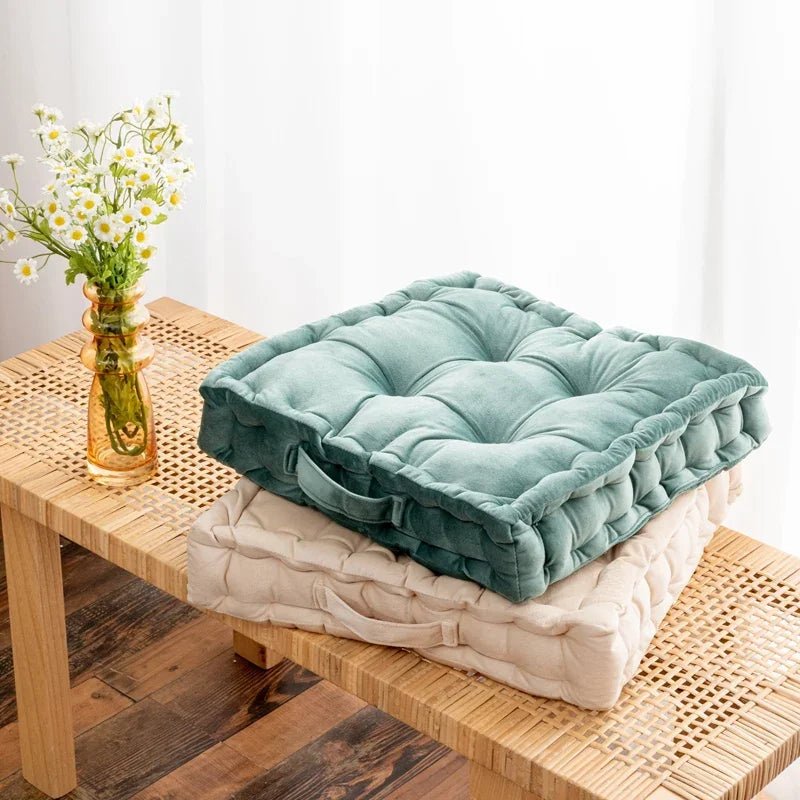 A teal square cotton throw pillow resting on a light beige cushion on a wooden table next to a vase of white flowers.