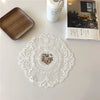 A white oval lace embroidered coaster is displayed on a white surface next to a wooden tray and a white container.