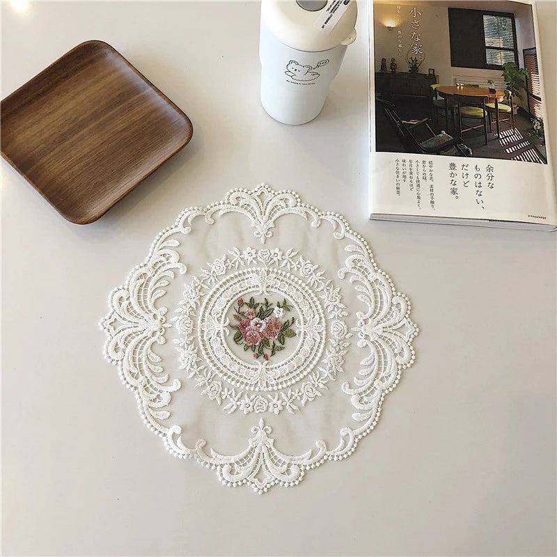 A white oval lace embroidered coaster is displayed on a white surface next to a wooden tray and a white container.