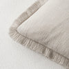 A beige linen throw pillow cover with a fringed edge resting on a white fabric background.