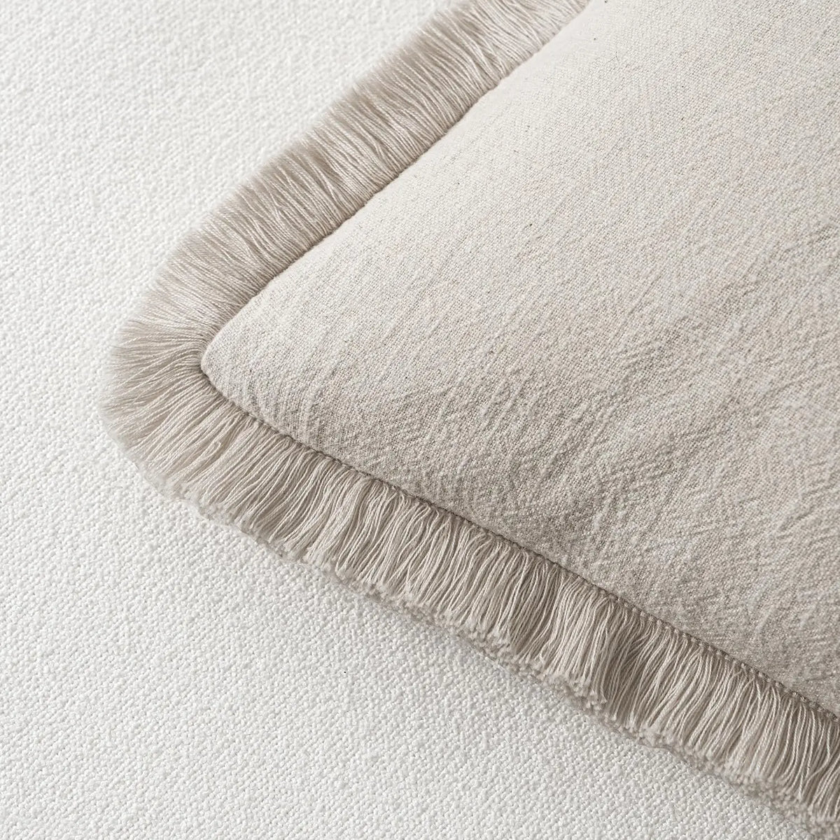 A beige linen throw pillow cover with a fringed edge resting on a white fabric background.
