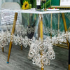A clear glass table is decorated with white lace embroidery and floral accents on a wooden surface.