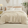 A beige quilted blanket is draped over a white duvet on a bed with a wooden headboard and natural light.