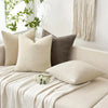 A beige linen textured throw pillow cover resting on a white sofa with a cream knitted throw blanket.