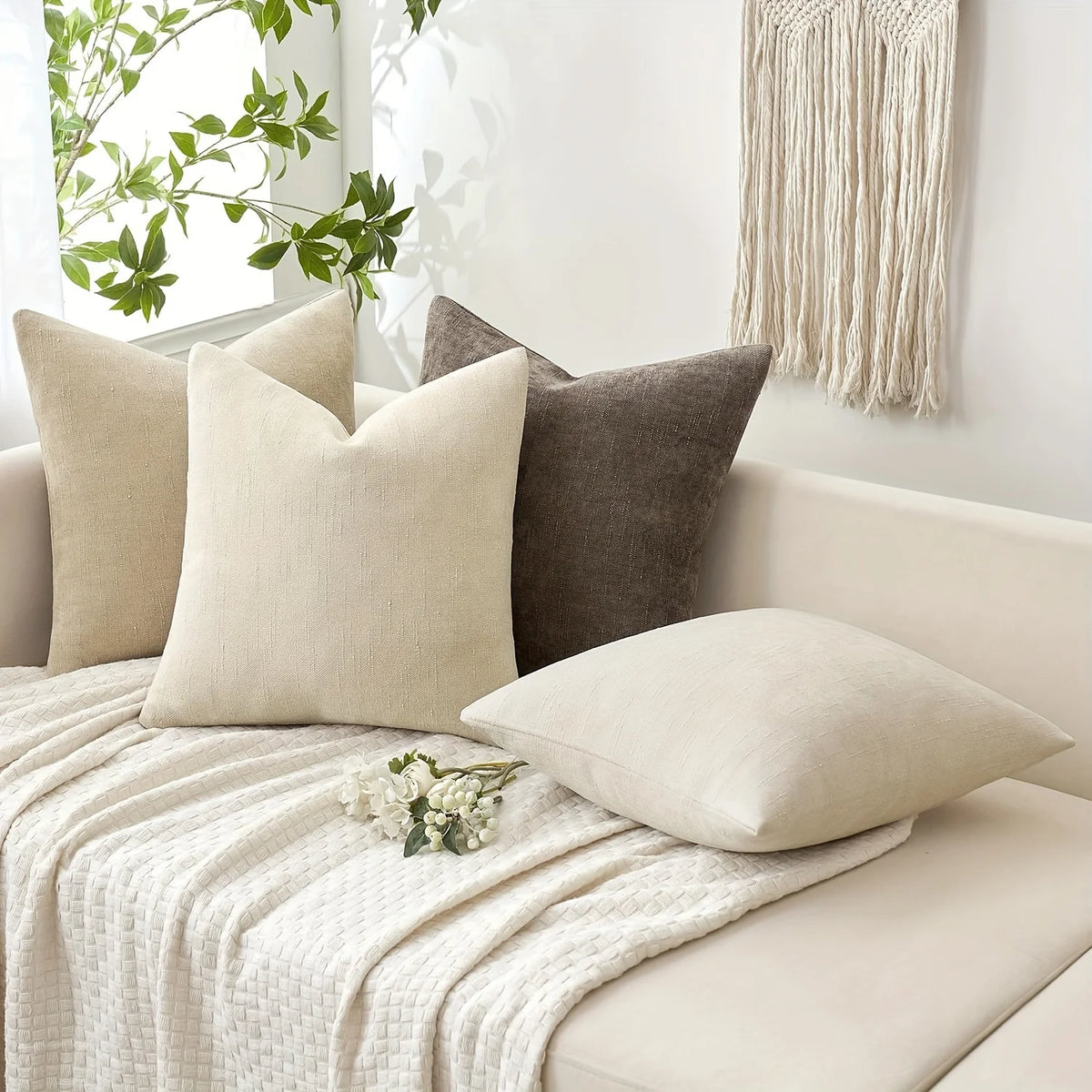 A beige linen textured throw pillow cover resting on a white sofa with a cream knitted throw blanket.