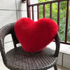 A bright red plush heart-shaped throw pillow made of fox fur resting on a dark brown wicker chair.