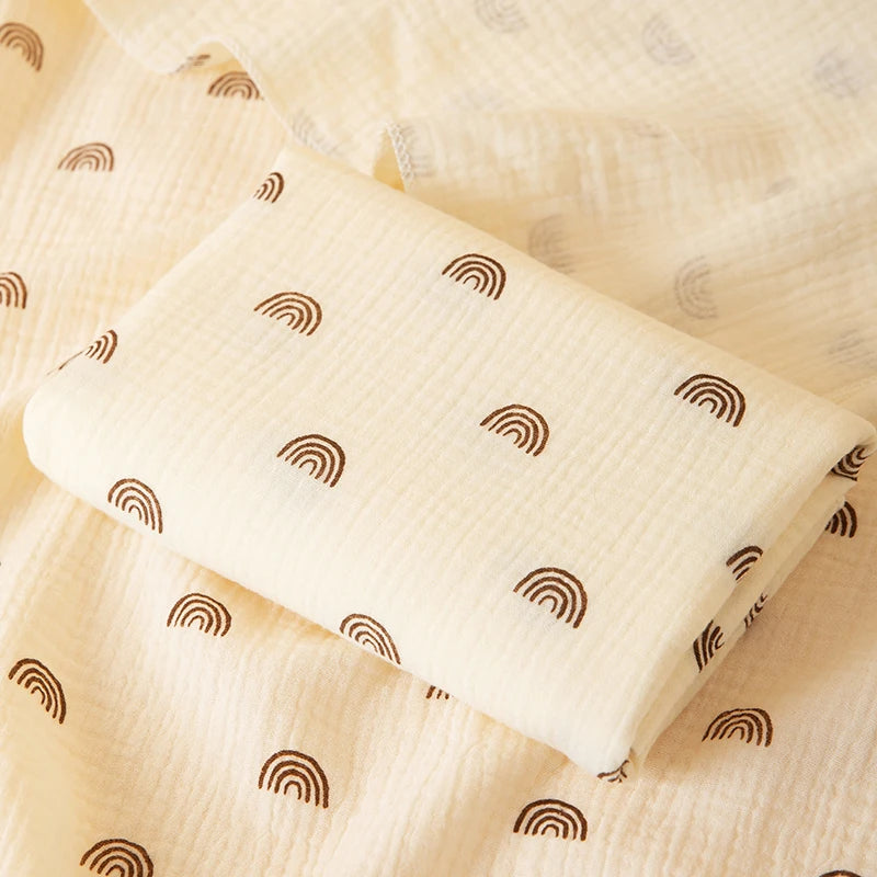 A cream-colored baby swaddle blanket with a subtle brown rainbow pattern is folded on a light fabric background.