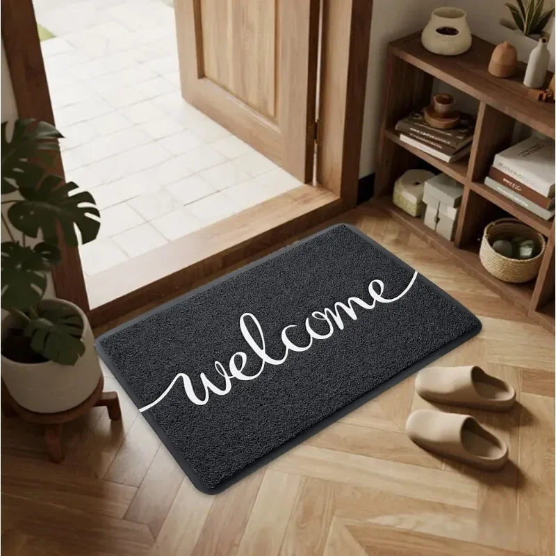 A dark gray minimalist script welcome doormat with white lettering placed on a light wood floor.