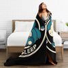 A woman is relaxing on a bed with a black and teal patterned throw blanket draped over her.