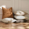 A stack of four linen textured throw pillows in shades of beige, tan, and gray on a wooden floor.