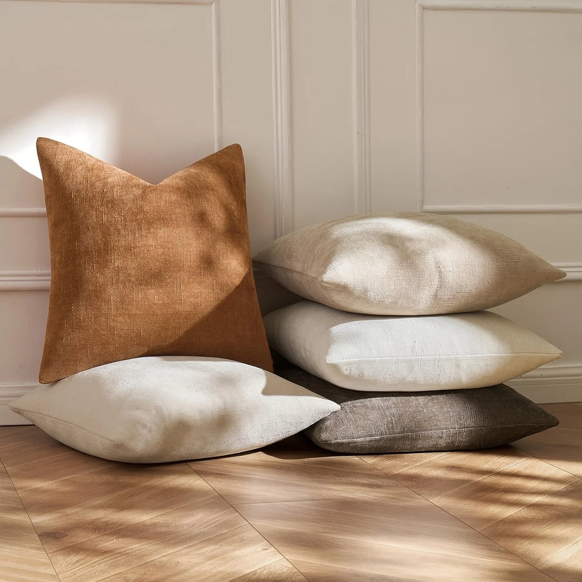 A stack of four linen textured throw pillows in shades of beige, tan, and gray on a wooden floor.