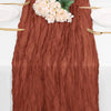 A rust colored cheesecloth table runner with a textured, wrinkled appearance, set for a rustic table with white plates and go
