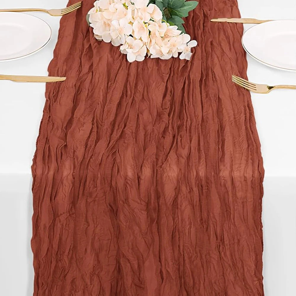 A rust colored cheesecloth table runner with a textured, wrinkled appearance, set for a rustic table with white plates and go