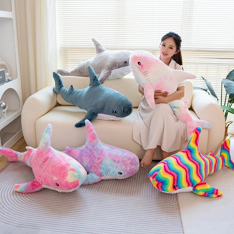 A woman is sitting on a couch surrounded by various giant shark plush toys in pink, blue, and gray colors.