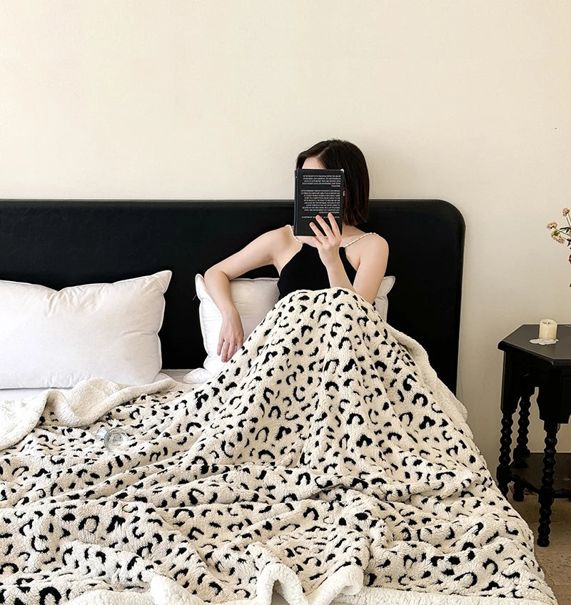 A woman is wrapped in a white and black leopard print jacquard knit throw blanket on a dark bed.
