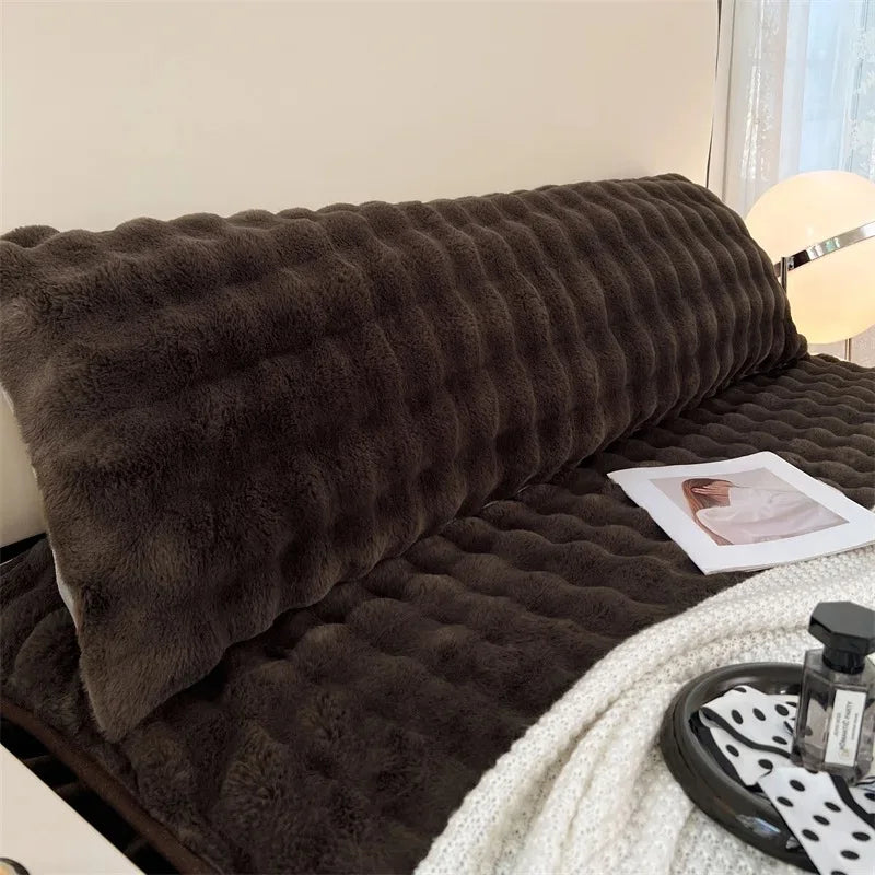 A dark brown plush long pillowcase resting on a bed, next to a white knitted blanket.