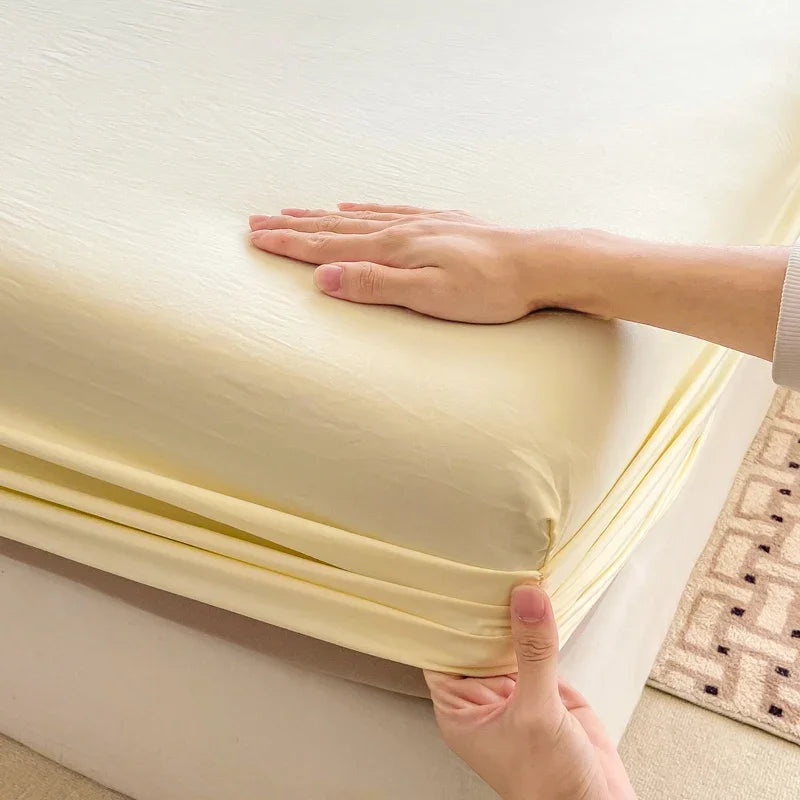 A hand is folding a light yellow fitted bed sheet on a white mattress.