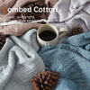 A close-up of a textured linen throw blanket in various shades of gray and blue, with a cup of coffee and pine cones.