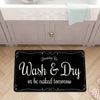 A black laundry mat with white text that reads Wash & Dry or be naked tomorrow placed on a wooden floor.