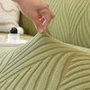 A hand is touching the textured, olive green fabric of a plant pattern stretch sofa cover.