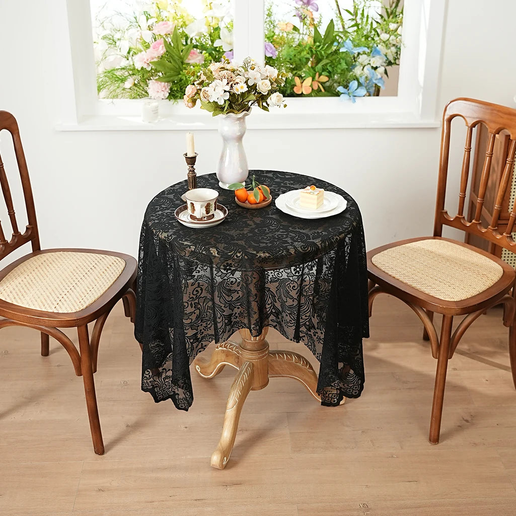 A black lace tablecloth is set on a round table with a vase of flowers and cups, surrounded by wooden chairs.