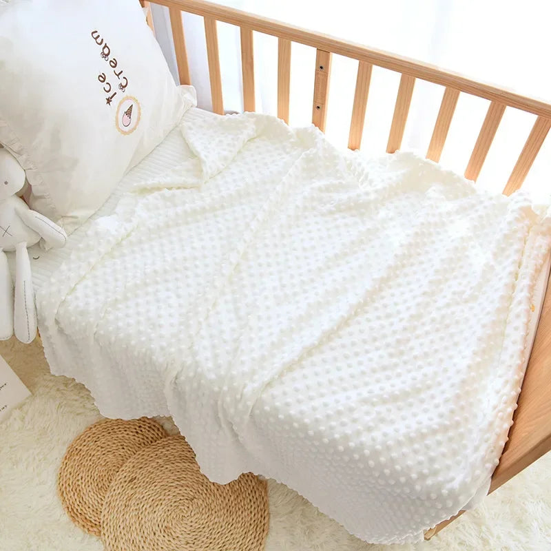 A white fleece swaddle blanket with a dotted pattern is displayed on a wooden crib railing.