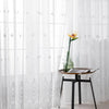 White sheer tulle window curtain with delicate silver floral embroidery and a vase holding an orange flower on a wooden table