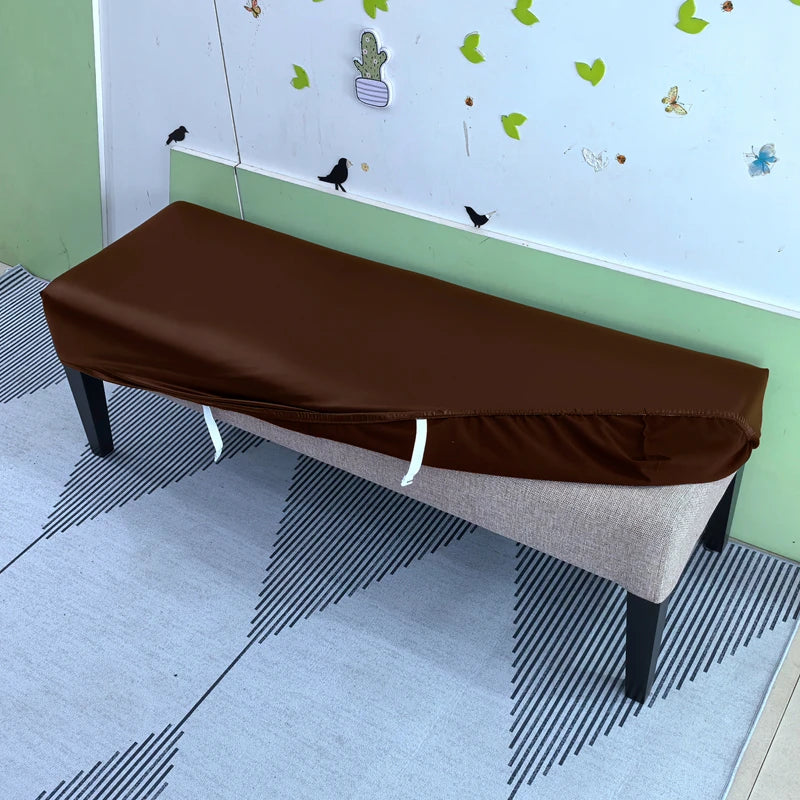 A brown stretch pu leather waterproof bench slipcover placed on a light gray floor in a room with a white wall and green trim