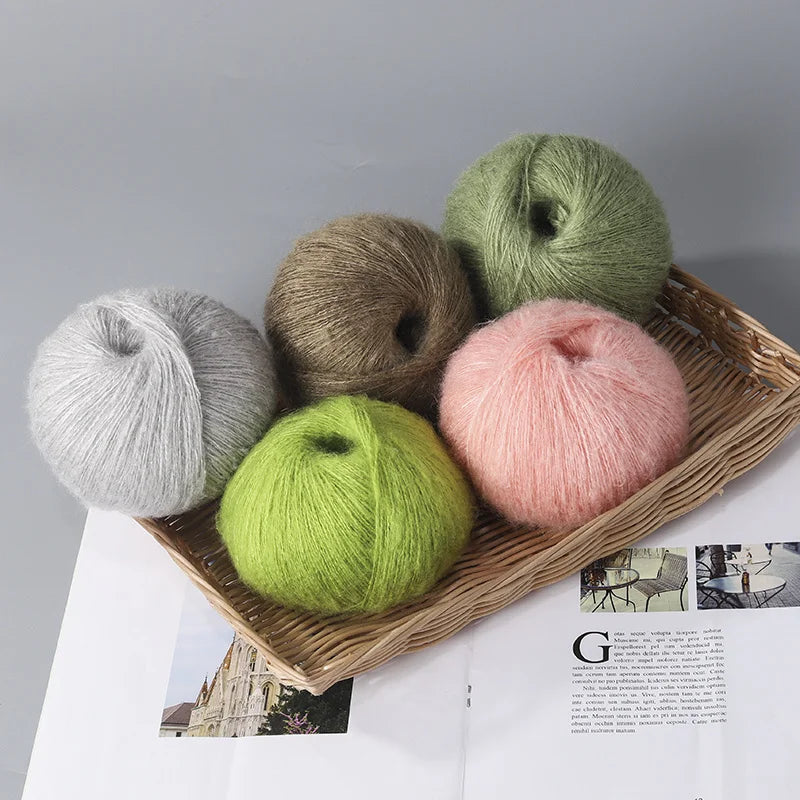 A collection of six balls of premium mohair yarn in various shades of green, pink, and gray arranged on a wooden tray.