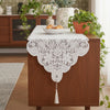 An embroidered white lace table runner is placed on a wooden cabinet with a bowl of bread and green foliage.