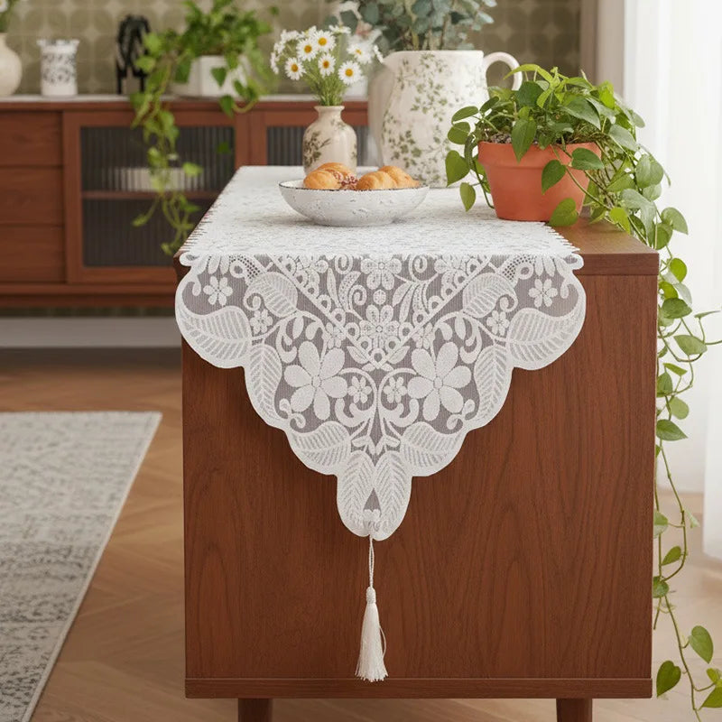 An embroidered white lace table runner is placed on a wooden cabinet with a bowl of bread and green foliage.