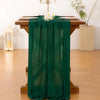 A dark green cheesecloth table runner draped over a wooden table with white flowers and gold candelabra.
