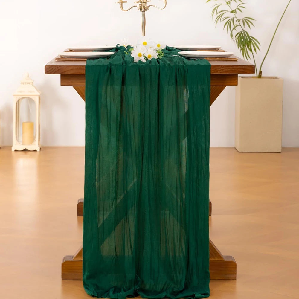 A dark green cheesecloth table runner draped over a wooden table with white flowers and gold candelabra.