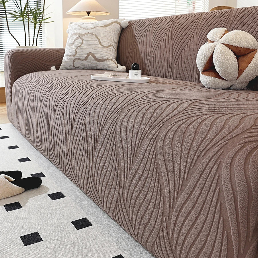 A close-up of a taupe stretch sofa cover featuring vertical ribbed texture and an elastic edge, displayed in a modern living 