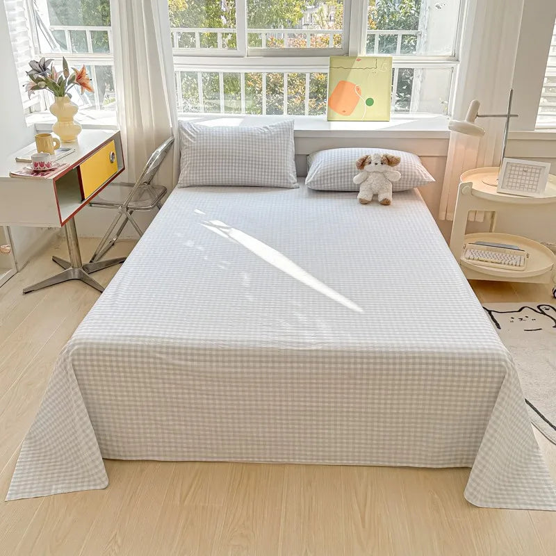 A white checkered flat sheet and pillow set is displayed on a bed in a bright room with large windows.