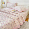 A pink floral quilted bedspread with matching pillows and a white wooden headboard.