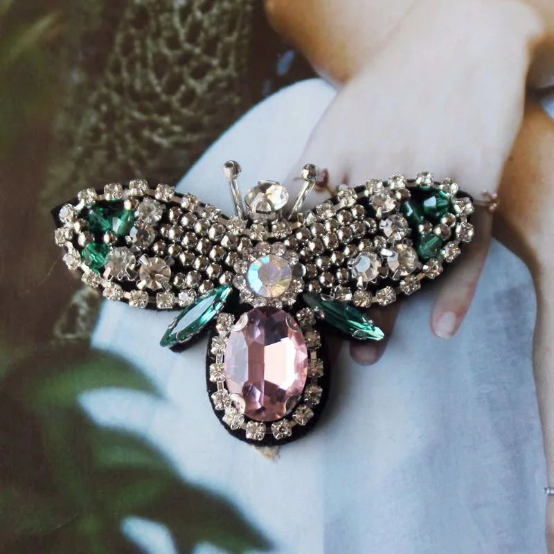 A hand holds a detailed handmade bee beaded sew-on patch featuring sparkling stones and green accents.