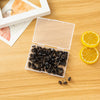 A clear plastic box containing a pack of round head push pins rests on a wooden surface next to a slice of lemon.