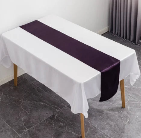 A white tablecloth with a deep purple satin table runner is set up on a table in an indoor setting.