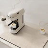 A white Mego espresso machine is placed on a beige kitchen drain mat on a white countertop.