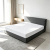 A modern bed with a gray upholstered headboard and white bedding sits on a light rug in a bright room.