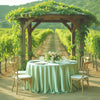 A lush vineyard setting featuring a wooden arbor with greenery and a round table draped in a sage green satin cloth.