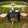 A black lace tablecloth is set on a round table outdoors with a white vase of flowers and small pastries.