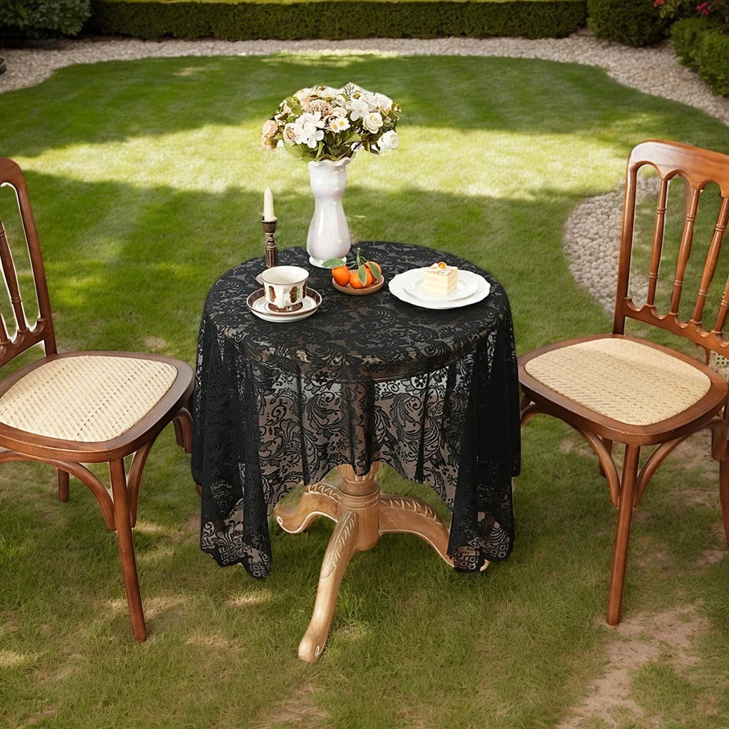 A black lace tablecloth is set on a round table outdoors with a white vase of flowers and small pastries.
