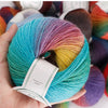 A hand holds a skein of rainbow wool yarn featuring various shades including blue, pink, yellow, and green.