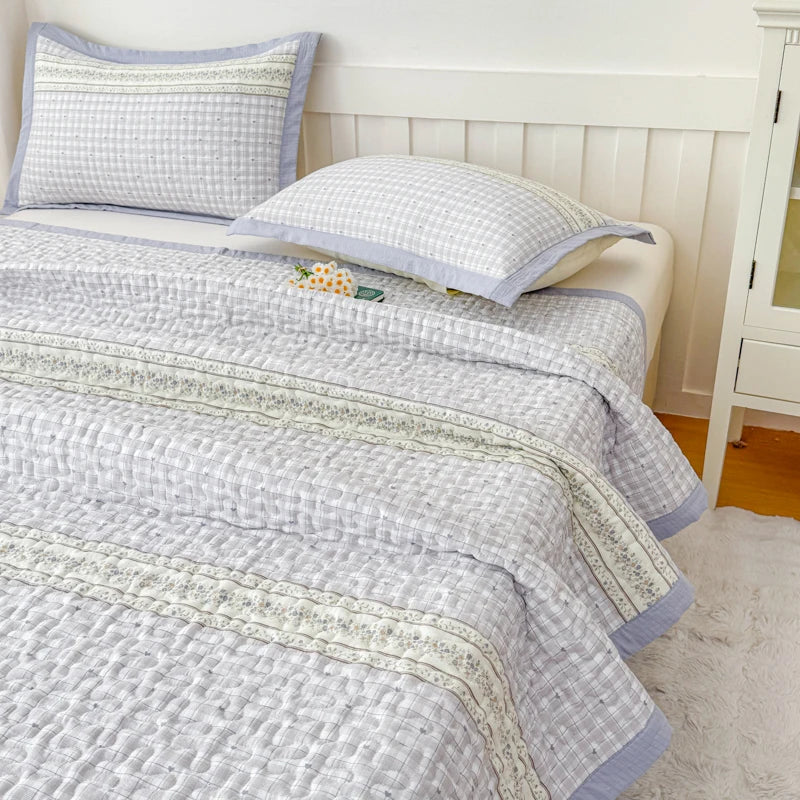 A light gray floral quilted bedspread with pale blue trim and pillows on a white bed.