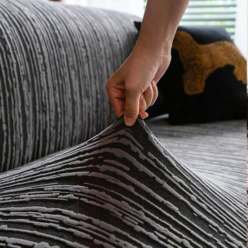 A hand is adjusting a dark gray and black striped jacquard plush elastic sofa cover on a sofa.