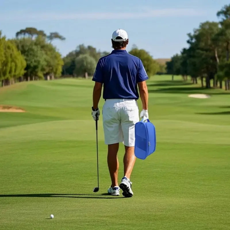 A man in a blue shirt and white shorts stands on a golf course, carrying a blue shoe carrier bag.