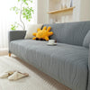 A gray stretchable sofa cover with a vertical line pattern is displayed on a light rug, featuring a yellow stuffed animal.