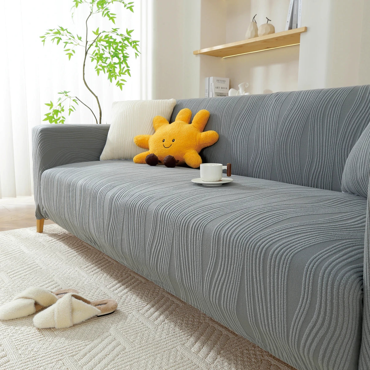 A gray stretchable sofa cover with a vertical line pattern is displayed on a light rug, featuring a yellow stuffed animal.